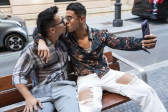 A latin gay couple shares a kiss while taking a selfie on a bench in Madrid. They display affection