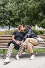 A loving LGBT multiethnic couple shares a tender kiss while sitting on a bench in a lush park. The