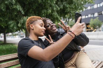 A joyful LGBT multiethnic couple taking a selfie on a park bench. They smile and flash peace signs,