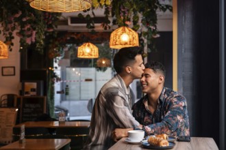A latin gay couple shares a tender kiss in a warmly lit cafe surrounded by lush decor. They enjoy