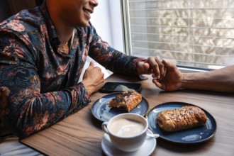 A latin gay couple shares a tender moment holding hands at a cafe. On the table are plates with