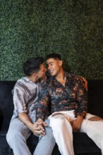 A loving latin gay couple sits closely on a sofa, holding hands and sharing a joyful, intimate