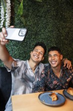 A joyful latin gay couple takes a selfie while enjoying brunch at a café. Their smiles and stylish