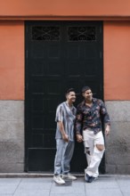 A joyful latin gay couple holds hands while standing in front of a colorful building in Madrid. The