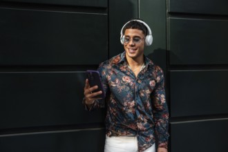 A fashionable man in sunglasses and a floral shirt stands against a dark green wall, wearing