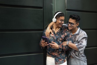 A joyful latin gay couple embraces while listening to music. One wears headphones and a floral
