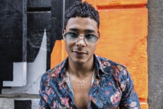 A confident latin man wearing sunglasses and a floral shirt poses against a vibrant, colorful