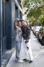 A happy latin gay couple taking a selfie on a bustling street in Madrid. They smile and embrace,