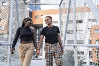 A fashionable LGBT multiethnic couple walks hand in hand across a modern footbridge, smiling and