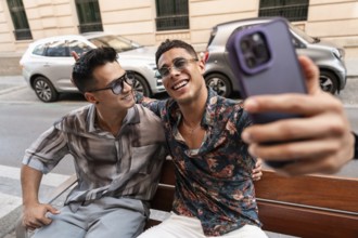 A joyful latin gay couple takes a selfie on a bench in Madrid. Both smiling, dressed in casual