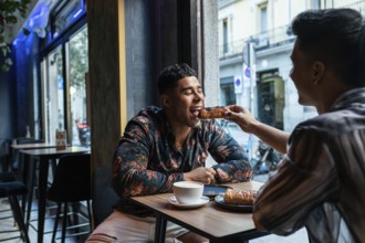 A joyful latin gay couple shares a pastry at a cafe, highlighting a moment of connection. The