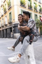 A joyful latin gay couple piggybacking on a picturesque street in Madrid. The colorful buildings