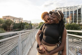 A happy LGBT multiethnic couple embraces and smiles warmly at each other on an urban bridge, with a