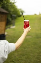 A hand holding a refreshing red drink with ice and a straw, set against a lush green background on