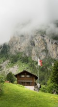A cozy wooden chalet stands amidst lush greenery, with misty cliffs in the background at Lac de