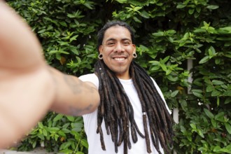 A cheerful man with long dreadlocks smiles while taking a selfie in a lush green garden in