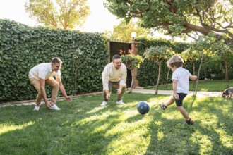A happy gay couple plays soccer with their young son in a sunlit garden. The lush greenery and