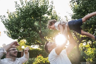 A happy gay couple plays with their young son outdoors on a sunny day, surrounded by lush greenery.