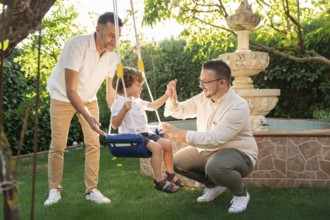 A joyful gay couple engages with their young son in a sunny garden, sharing laughter and high-fives