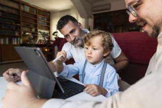 A gay couple sits with their young son in a cozy living room, focused on a tablet. They are sharing