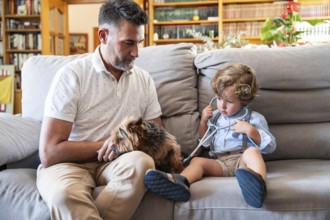A boy with a stethoscope playfully examines a dog while sitting on a couch. His grandfather watches
