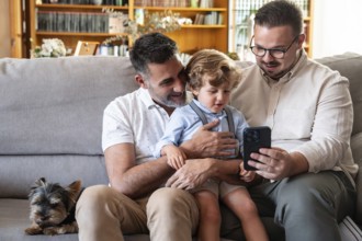 A joyful gay couple sits on a cozy couch with their young son and a small dog, sharing moments