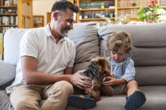A joyful father and his young son play doctor and patient with their pet dog on a comfortable