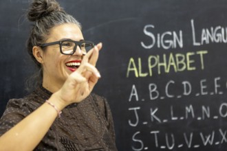 A woman teaches sign language alphabet in a classroom, gesturing with a smile Chalkboard filled