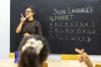 A sign language instructor demonstrates letters from the alphabet on a blackboard in a classroom