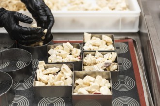 A baker wearing black gloves carefully arranges small pieces of dough in square molds on a baking