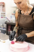 A skilled artisan wearing an apron and gloves adds delicate floral decorations to a pink pastry,