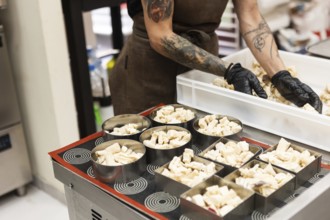 A focused baker with tattooed arms and black gloves arranges pastry pieces into metal molds,