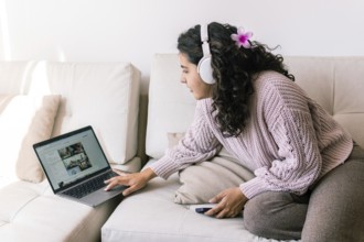 A young latin woman relaxes on a cozy sofa at home, wearing headphones and browsing her laptop She