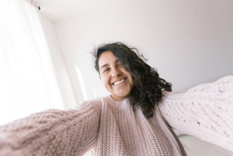 A cheerful young latin woman with curly hair wears a cozy pink sweater, smiling as she enjoys a