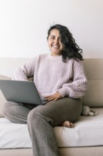 A cheerful young hispanic woman sits comfortably on a sofa at home, using her laptop She is dressed