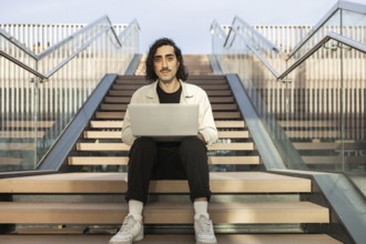 A man sits on an outdoor stairway, focused on his laptop Dressed casually, he embodies a modern,