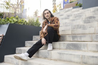 A man sits on outdoor steps, showcasing a relaxed, casual urban style Wearing a patterned jacket,