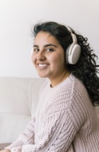 A young latin woman sits on a cozy couch, wearing headphones and a knitted sweater She is smiling,