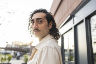 A stylish young man with curly hair and a mustache stands confidently outdoors, backlit by the