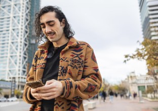 A fashionable man with curly hair wearing a patterned jacket checks his smartphone on a city street