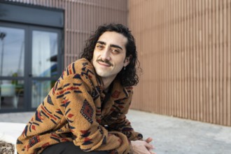 A man with long hair and a mustache, wearing a cozy patterned jacket, smiles while sitting outdoors