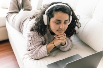 A young latin woman relaxes on a couch, wearing headphones and a cozy sweater, as she engages with