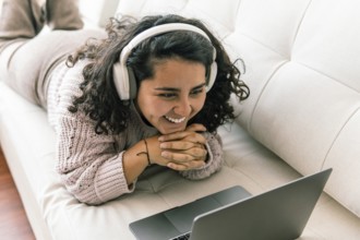 A young latin woman wearing headphones lies on a sofa, smiling warmly as she watches something on