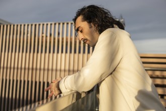 A man in a light shirt leans on a railing, captured in soft evening light His thoughtful expression