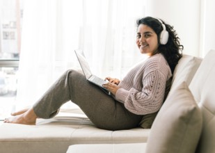 A young latin woman in cozy attire sits comfortably on a sofa at home, using a laptop with