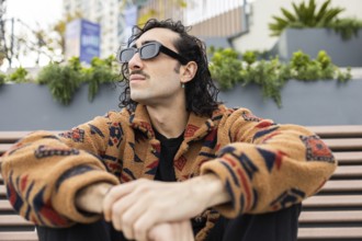 A fashionable man wearing sunglasses and a patterned jacket sits on a bench outdoors, exuding a