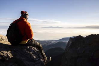 Back view of unrecognizable man sitting atop a mountain during a breathtaking sunrise, equipped