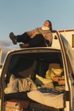 A Latina woman laughs joyfully as she sits in the open trunk of a car, surrounded by luggage and