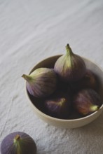 A rustic ceramic bowl filled with ripe, purple figs placed on a textured linen tablecloth,