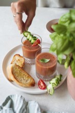 A hand garnishes gazpacho with cucumber and tomato skewers in two glass cups The plate also has